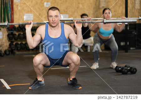 Motivated man practicing exercises with rod near other people in gym 142787219