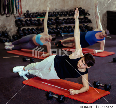 Energetic young man doing side-plank near other people in gym Energetic young man doing side-plank near other people in gym 142787372
