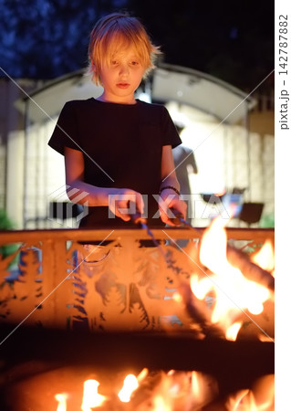 A cute boy is roasting sausages using skewers over a campfire on a summer evening. An active outdoor getaway for a family with children during a countryside vacation. Camping A cute boy is roasting sausages using skewers over a campfire on a summer evening. An active outdoor getaway for a family with children during a countryside vacation. Camping 142787882