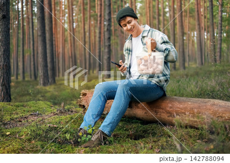 man sitting on a forest log holding the smartphone and a basket full of wild mushrooms, enjoying a seasonal autumn harvest, foraging, and nature connection. man sitting on a forest log holding the smartphone and a basket full of wild mushrooms, enjoying a seasonal autumn harvest, foraging, and nature connection. 142788094