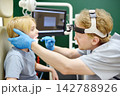 An experienced otolaryngologist examines a patient's throat using a medical tongue depressor in the consulting room. The professional ENT specialist is at their workplace. 142788926