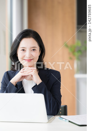 ノートパソコンで仕事をする女性会社員、オフィスで笑顔で働くビジネスウーマンのデスクワークカメラ目線 ノートパソコンで仕事をする女性会社員、オフィスで笑顔で働くビジネスウーマンのデスクワークカメラ目線 142789685