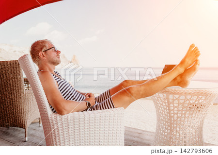 Man relaxing beach sits in a white wicker chair with his feet up on a table at a coastal resort terrace under the sun 142793606