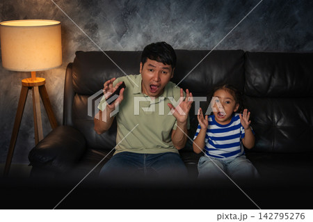 shocked and scared father and daughter watching horror movie tv in the living room at night shocked and scared father and daughter watching horror movie tv in the living room at night 142795276