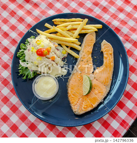 Fish steak with vegetables, French fries, sauce on a blue plate, placed on a checkered tablecloth. Fish steak with vegetables, French fries, sauce on a blue plate, placed on a checkered tablecloth. 142796336