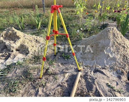 A surveying tripod standing between piles of sand in the garden. site preparation, land surveying,garden renovation. A surveying tripod standing between piles of sand in the garden. site preparation, land surveying,garden renovation. 142796903