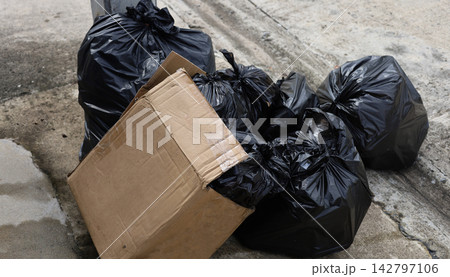 Black plastic garbage bags piled up next to a discarded cardboard box on a concrete sidewalk. Black plastic garbage bags piled up next to a discarded cardboard box on a concrete sidewalk. 142797106