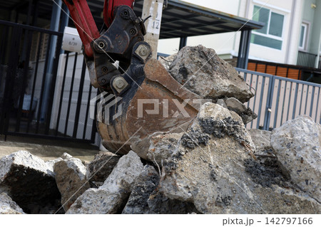 Close-up of an excavator bucket scooping heavy concrete pieces from a pile at a construction site. Close-up of an excavator bucket scooping heavy concrete pieces from a pile at a construction site. 142797166