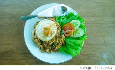 Indonesian traditional fried rice or Nasi Goreng served on a white plate with a sunny-side-up egg, fresh lettuce, sliced cucumber, and tomato garnish, placed on a wooden table background. Top view, hi Indonesian traditional fried rice or Nasi Goreng served on a white plate with a sunny-side-up egg, fresh lettuce, sliced cucumber, and tomato garnish, placed on a wooden table background. Top view, hi 142798003