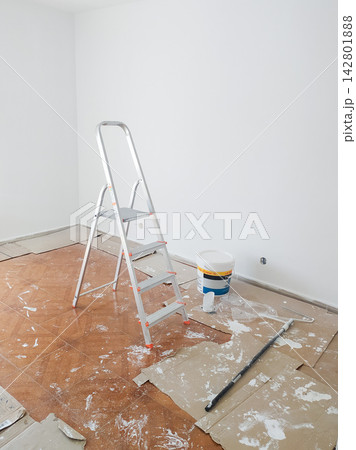 A metal stepladder and paint bucket stand in an empty room during a home renovation. White splatters on parquet floor symbolize DIY improvement, creativity, and a fresh start. A metal stepladder and paint bucket stand in an empty room during a home renovation. White splatters on parquet floor symbolize DIY improvement, creativity, and a fresh start. 142801888