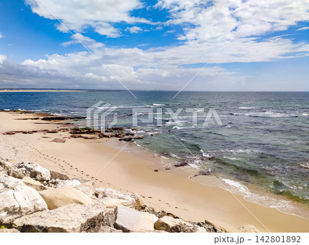 This serene coastal landscape features a pristine sandy beach, rugged rocks, and gentle turquoise ocean waves under a bright, cloudy blue sky, evoking peaceful solitude. Nobody. 142801892