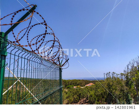A green security fence topped with rusty razor wire stands against a clear blue sky and lush forest, evoking confinement. Nobody is present in this powerful metaphor for safety. A green security fence topped with rusty razor wire stands against a clear blue sky and lush forest, evoking confinement. Nobody is present in this powerful metaphor for safety. 142801961