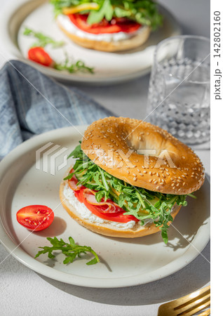 Two fresh bagels with tomato, prosciutto, cheese and arugula on a plates on a light background Two fresh bagels with tomato, prosciutto, cheese and arugula on a plates on a light background 142802160