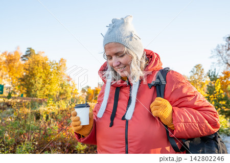 Woman hiker in autumn forest holding disposable paper coffee cup. Mature forty seven year old lady relaxing during trekking trip. Bodypositive active lifestyle in outdoor environment. 142802246