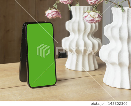 Smartphone mockup with a blank white screen leans on a wooden table near decorative ceramic vases Smartphone mockup with a blank white screen leans on a wooden table near decorative ceramic vases 142802303