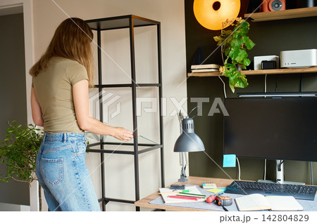 Woman placing glass shelf into shelving unit beside home office desk 142804829