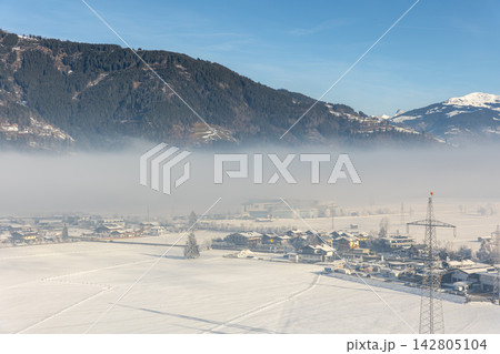 Snowy Kaprun valley lies beneath forested Austrian Alps, wrapped in low winter fog and bright sunlight. Power pylons cross frozen fields near village, roads, farms, and industrial buildings 142805104