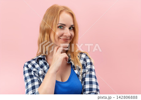 Young woman with red hair and blue eyes looking at the viewer with a clever smirk, her hand resting under chin, conveying a sense of intrigue, secret, or thoughtful consideration 142806888