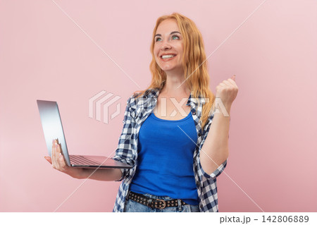 Joyful woman holding a notebook computer and making a winning gesture, experiencing online achievement or conceptualizing a new idea, isolated on a pink background Joyful woman holding a notebook computer and making a winning gesture, experiencing online achievement or conceptualizing a new idea, isolated on a pink background 142806889