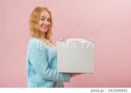 Smiling red haired woman holding a blank white cardboard carry box with a handle, presenting a delivery or a gift against a pink background Smiling red haired woman holding a blank white cardboard carry box with a handle, presenting a delivery or a gift against a pink background 142806891