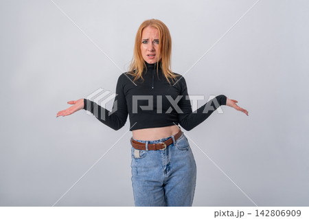 Young woman shrugging with open hands and puzzled expression, conveying confusion, doubt, or asking why while standing against a plain studio background 142806909