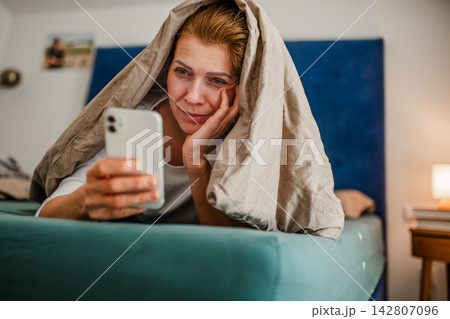 Woman lying under blanket and scrolling on phone. Woman lying under blanket and scrolling on phone. 142807096