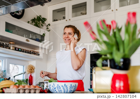 Mature woman making phone call while cooking in kitchen. Mature woman making phone call while cooking in kitchen. 142807104