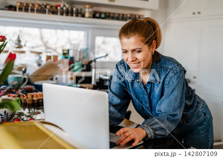 Mature woman searching recipe online on laptop while cooking in kitchen. Mature woman searching recipe online on laptop while cooking in kitchen. 142807109