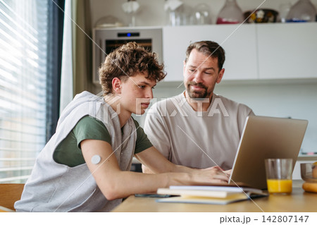 Father helping teenage son with diabetes with homework on laptop in kitchen. Father helping teenage son with diabetes with homework on laptop in kitchen. 142807147