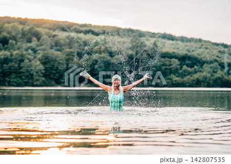 Cold water swimming for elderly women. Senior sporty women splashing in the water, standing in lake during cold evening, arms in the air. Cold water swimming for elderly women. Senior sporty women splashing in the water, standing in lake during cold evening, arms in the air. 142807535