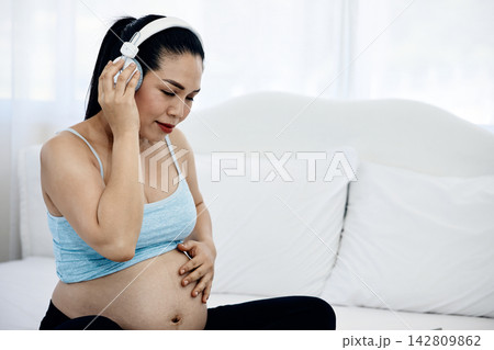 Pregnant woman wearing headphones while touching belly on bed, listening to music with calm expression for maternity wellness, relaxation, pregnancy care and peaceful bonding Pregnant woman wearing headphones while touching belly on bed, listening to music with calm expression for maternity wellness, relaxation, pregnancy care and peaceful bonding 142809862