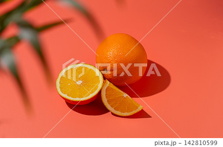 On the coral surface, next to two neatly sliced slices, there is a whole orange. Sunlight casts soft shadows, emphasizing the freshness and bright color of the fruit. 142810599