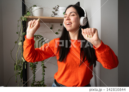 Young woman dancing with headphones beside houseplants in a bright home interior. Music therapy emotional wellbeing and self care 142810844