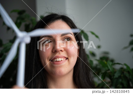 Smiling woman looks past a small wind turbine model in a leafy indoor setting. Clean energy renewable power and female representation Smiling woman looks past a small wind turbine model in a leafy indoor setting. Clean energy renewable power and female representation 142810845