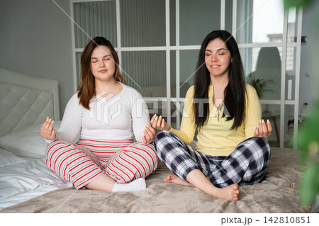 Two female friends meditate together on a bed in a bright home bedroom wearing comfortable sleepwear. Mindfulness wellbeing friendship and relaxation Two female friends meditate together on a bed in a bright home bedroom wearing comfortable sleepwear. Mindfulness wellbeing friendship and relaxation 142810851