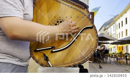 Male fingers of musician playing the bandura on a sunny summer day. Sunlight falls on the strings and wood surface, creating a warm and lively scene of outdoor music and authentic street performance Male fingers of musician playing the bandura on a sunny summer day. Sunlight falls on the strings and wood surface, creating a warm and lively scene of outdoor music and authentic street performance 142810933