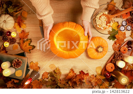 woman preparing for Halloween by carving a pumpkin lantern at home. Getting ready for an autumn holiday with hollowed squash, seeds, leaves and glowing candles on table. 142813622