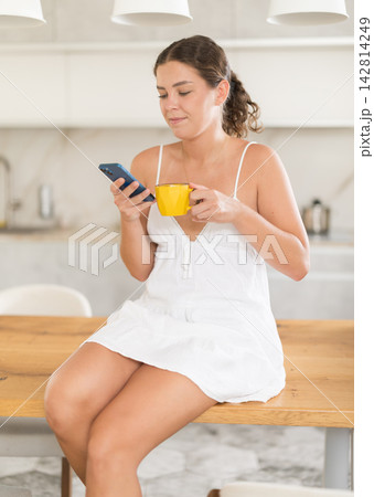 Girl with smartphone drinks coffee in kitchen Girl with smartphone drinks coffee in kitchen 142814249