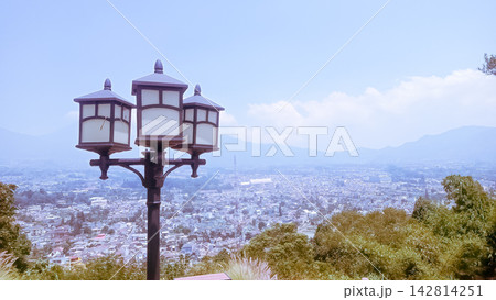 A vintage three-headed street lamp post standing on a high hill overlooking a scenic panoramic city view under a clear blue sky with distant mountains. 142814251