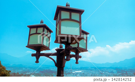 A vintage three-headed street lamp post standing on a high hill overlooking a scenic panoramic city view under a clear blue sky with distant mountains. 142814252