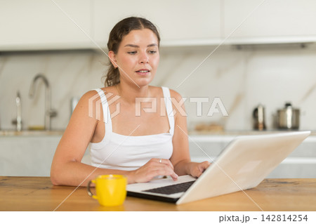 Portrait of young girl or housewife wearing home jersey and shorts working at laptop sitting at kitchen, remote work, freelance Portrait of young girl or housewife wearing home jersey and shorts working at laptop sitting at kitchen, remote work, freelance 142814254