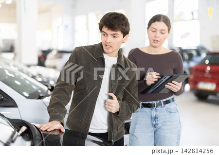 Young couple is standing in a car dealership and looking at the car with gadget in hands 142814256