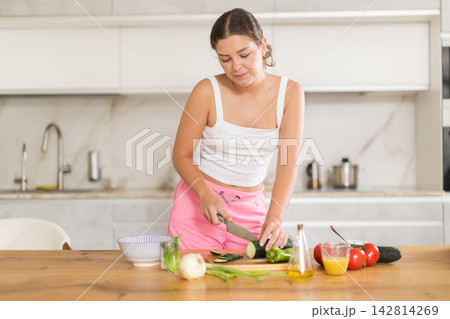 Happy young girl cutting fresh vegetables for salad on kitchen table Happy young girl cutting fresh vegetables for salad on kitchen table 142814269