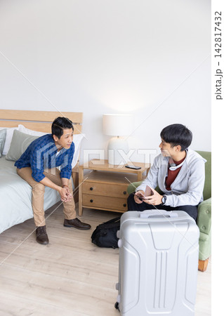 Two Asian men sitting in hotel room with silver suitcase, one wearing gray hoodie holding passport 142817432