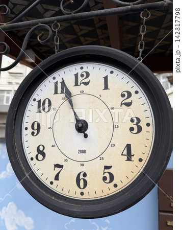 Round wall clock shows time at 10:10 with a black frame, hanging on a decorative structure outdoors against a blue background 142817798