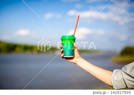 Hand holds a green and blue disposable cup with a straw, set against a serene river landscape and clear blue sky in the background Hand holds a green and blue disposable cup with a straw, set against a serene river landscape and clear blue sky in the background 142817939