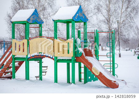 Colorful playground equipment with a slide and climbing structure is covered in snow, set in a winter park with trees in the background 142818059