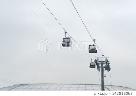Cable cars travel along overhead lines in an urban area, with a large building structure visible beneath them against a cloudy sky Cable cars travel along overhead lines in an urban area, with a large building structure visible beneath them against a cloudy sky 142818068