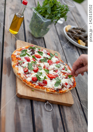 Freshly baked pizza with cherry tomatoes and basil on a wooden cutting board, accompanied by olive oil and herbs in the background 142818502
