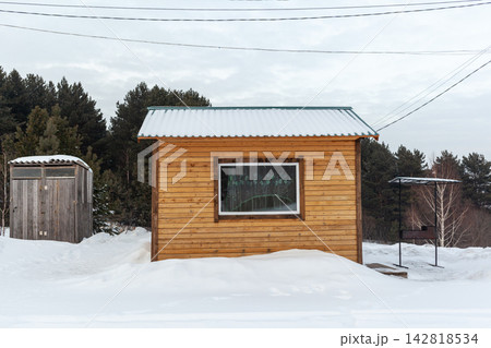 Wooden cabin surrounded by snow with a forest backdrop and an outdoor shed, power lines overhead in a winter landscape Wooden cabin surrounded by snow with a forest backdrop and an outdoor shed, power lines overhead in a winter landscape 142818534